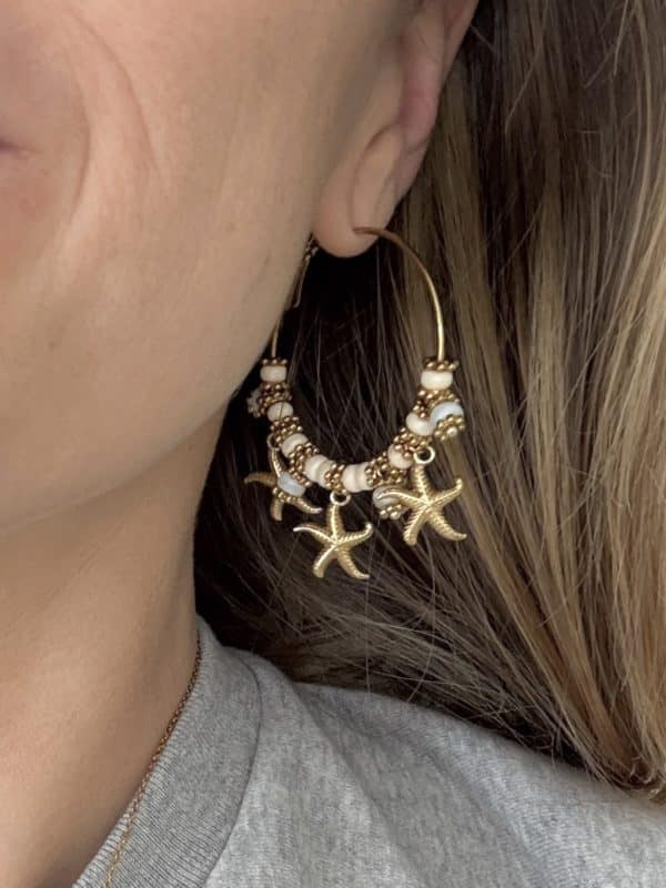 Boucles d’oreilles en acier inoxydable de couleur or, composées de deux créoles disposant chacune de trois pendentifs en forme d'étoile de mer. Vous y trouverez également des perles de coquillage et de céramique blanche